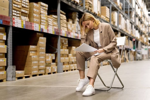 Stressed woman reviewing documents in a warehouse while seated on a folding chair.
